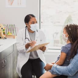 With a protective mask on, a female pediatrician talks to a young patient's mother about the woman's daughter's medical conditions. They are wearing protective masks during an office visit