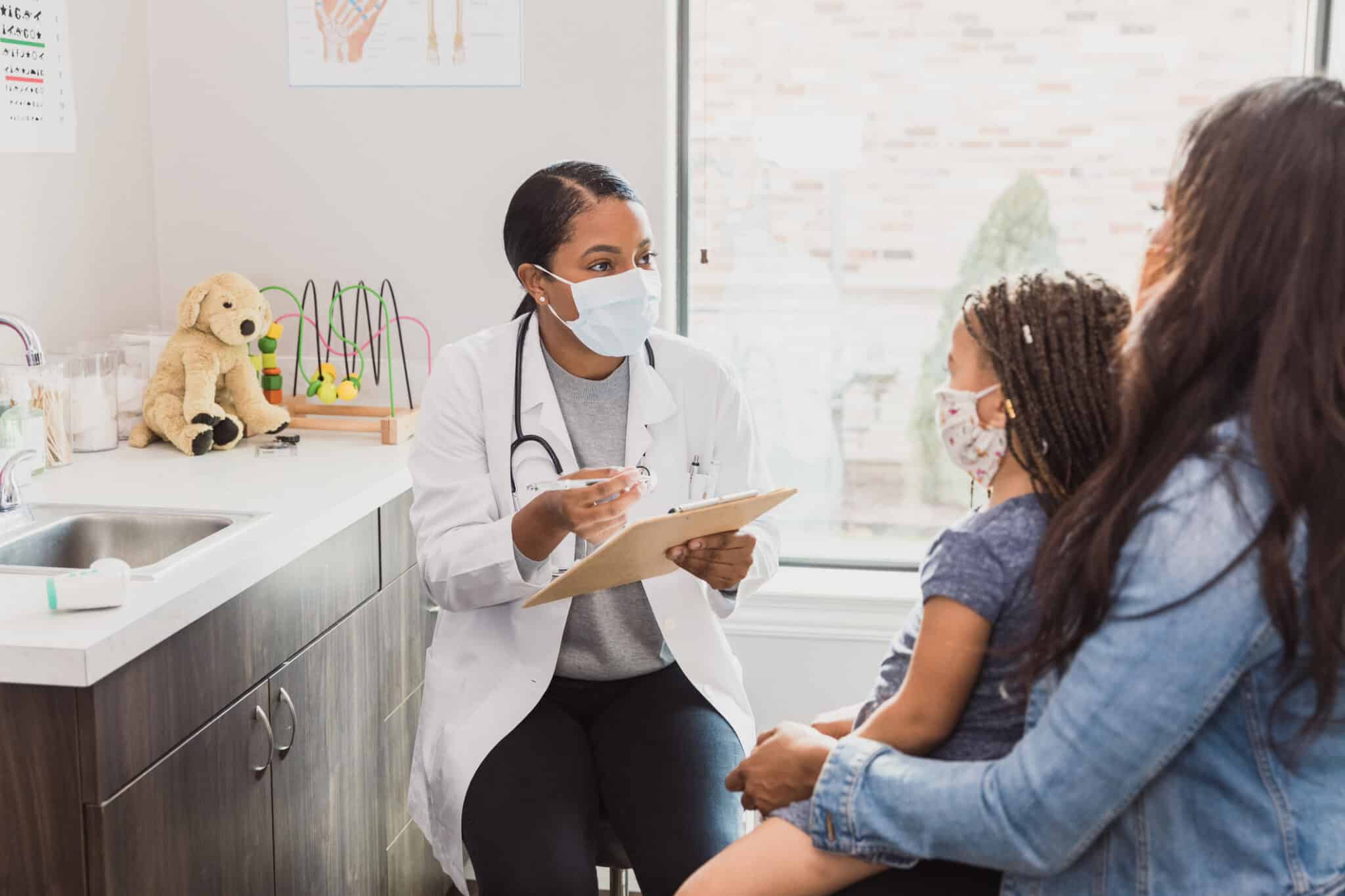 With a protective mask on, a female pediatrician talks to a young patient's mother about the woman's daughter's medical conditions. They are wearing protective masks during an office visit